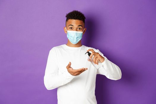 Concept of covid-19, pandemic and social distancing. Image of young african-american guy using hand sanitizer, wearing medical mask and white sweatshirt, standing over purple background.