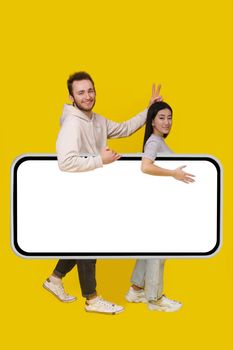 Carrying huge smartphone with white screen handsome guy set horns gesture on asian girl, mobile app advertisement and excited smile on camera isolated on yellow background. Product placement.
