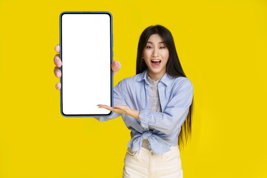 Big smartphone with white screen in hand of beautiful asian girl happy to introduce new app, game, win, isolated on yellow background. Mobile app advertisement product placement.