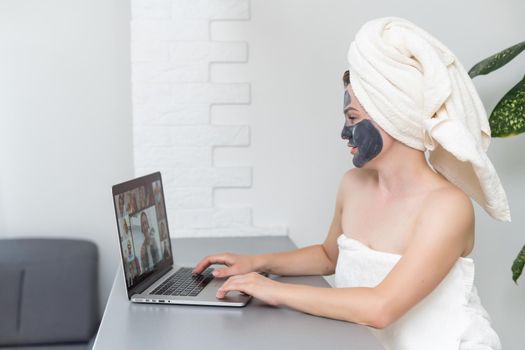 Young woman with towel on head and black facial clay mask sitting at home and using laptop computer . Staying at home, quarantine, distance learning, online education, technology, modern lifestyle.