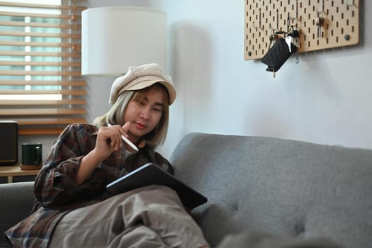 Asian woman working with digital tablet while sitting on couch.