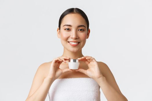 Beauty, personal care, spa salon and skincare concept. Tender beautiful asian girl in bath towel showing cream and smiling, recommend moisturizing, hydration product, white background.