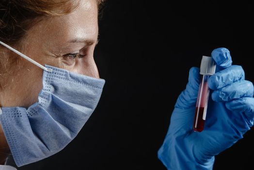 the doctor is holding a test tube of blood against a black background. A woman nurse in a mask is holding a flask of blood. Medical worker in protective mask working with preparations on black background. Close up