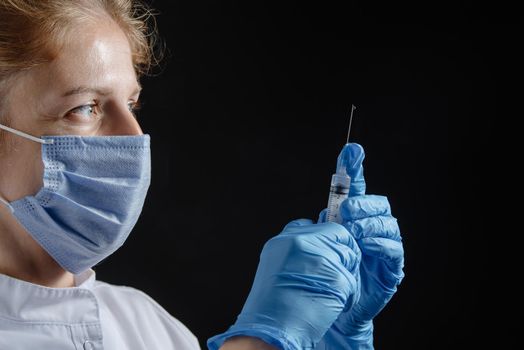 A female doctor wearing a protective mask and gloves holds a syringe with a vaccine or injection against a black background. A female nurse prepares to give an injection against a dark background. Woman with syringe in hand alone on black background. A drop of liquid on the syringe.