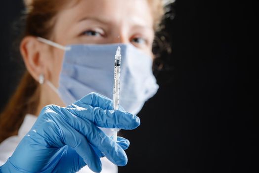 A young female doctor with a syringe looks at the camera. A nurse wearing a medical mask and blue latex gloves. Medicine and health care. Vaccination for the virus. Preventive measures against the disease. A place to copy.