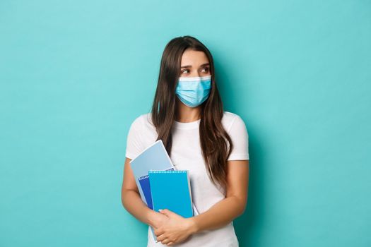 Concept of coronavirus, health and social distancing. Portrait of female student attend classes in medical mask, looking right at copy space, holding notebooks, standing over blue background.