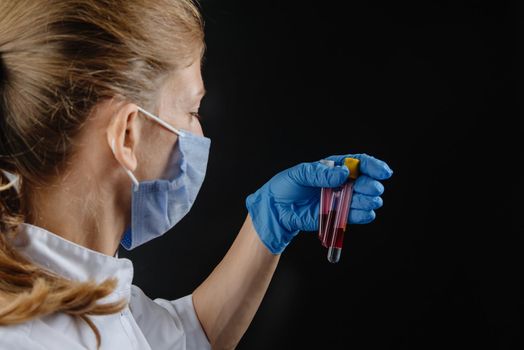 doctor holding blood vials against a black background. A woman nurse in a mask holds flasks of blood. Young doctor on black background turned away from camera. Medical worker in protective mask working with preparations on black background.