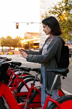 vertical photo of a young girl taking a bike with her smart phone in a public parking of rental bicycles in the city, concept of active lifestyle and sustainable mobility