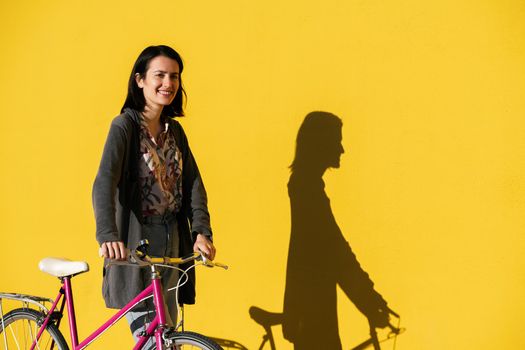 smiling young woman walking in front of a colorful yellow wall with her pink retro bike, concept of active lifestyle and sustainable mobility, copy space for text