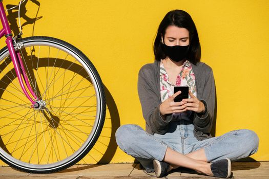 young girl wearing a protective mask consulting her smart phone sitting next to her bike leaning against a yellow wall, concept of technology, protection against covid and sustainable mobility