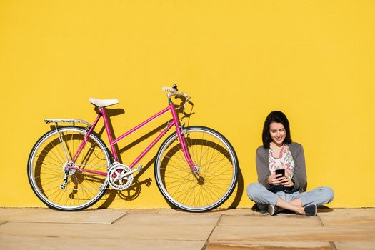 happy young girl looking smart phone sitting next to her bike leaning against a colorful yellow wall, concept of technology and sustainable mobility, copy space for text