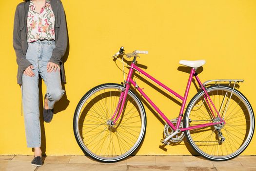 unrecognizable young woman leaning against a colorful yellow wall next to her pink retro bike, concept of active lifestyle and sustainable mobility, copy space for text