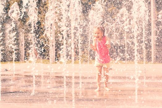 excited baby girl having fun between water jets, in fountain. Summer in the city