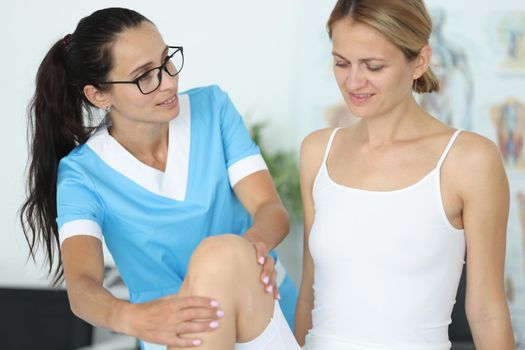 Female patient receiving medical care in rehabilitation center after leg injury. Physiotherapist examining leg and knee of woman sitting on medical couch