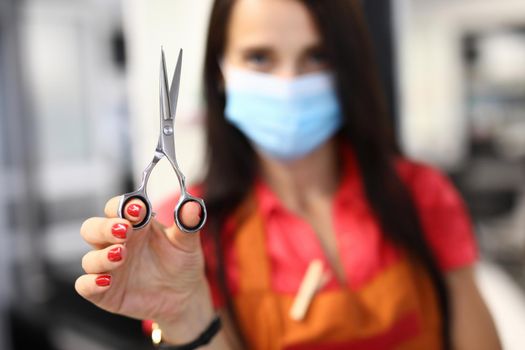 Woman hairdresser in protective mask holds scissors. Health safety and provision of services in beauty salon