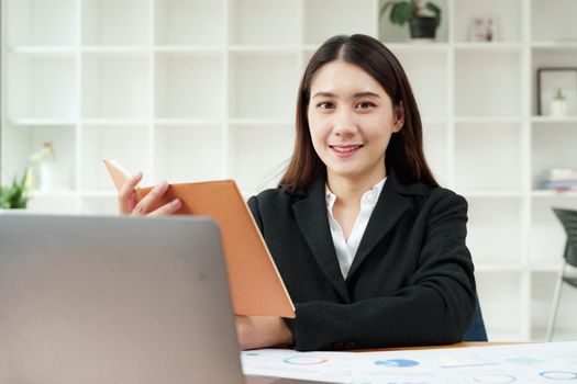 Portrait of a beautiful Asian businesswoman in a formal suit sits at her office desk with using notebook and laptop computer in office.