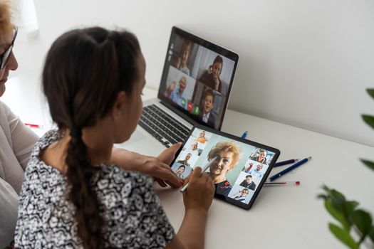 Portrait of happy grandmother and little granddaughter making video conference on pc sitting at table, waving hands at screen, greeting somebody, chatting with parents, enjoying online communication