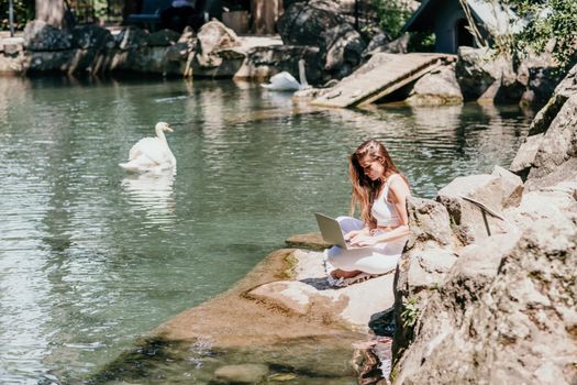 a beautiful woman in a long white dress sits near a lake with swans and works at a laptop.