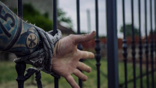 Details of tattooed hands of a man tied to grills behind him struggles to escape