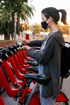 vertical photo of a young girl with protective mask cleaning her hands with disinfectant gel next to an urban bicycle rental parking, concept of protection against covid and sustainable mobility