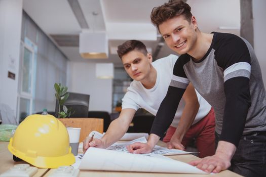 Success, career, startup concept. Handsome cheerful male architect smiling to the camera, while working on blueprints with his colleague