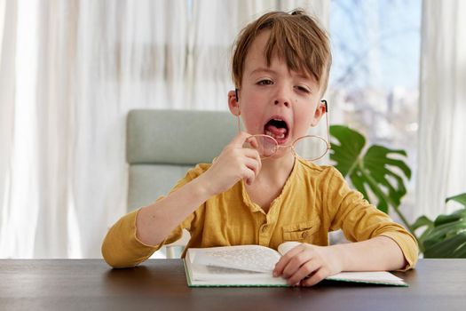Little boy in yellow shirt taking off glasses and yawning while sitting at table with book and doing homework in daytime at home