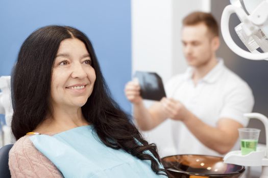 Oral checkups. Beautiful senior woman smiling looking away sitting in a dental chair at the clinic her dentist examining teeth x-ray on the background patient people medicine healthcare smile concept
