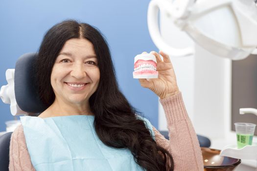 Her smile is perfect. Beautiful happy senior woman sitting relaxed in dental chair smiling showing dental teeth mold to the camera copyspace dentistry dental clinic patient healthcare medicine concept