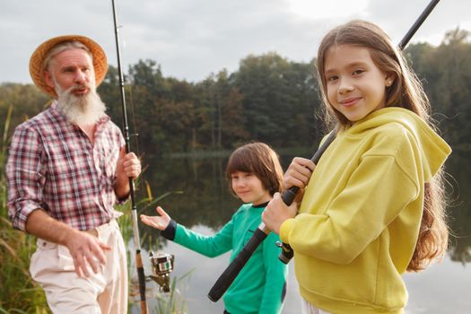 Cute happy young girl smiling to the camera, holding fishing rod, her grandpa and brother on the background