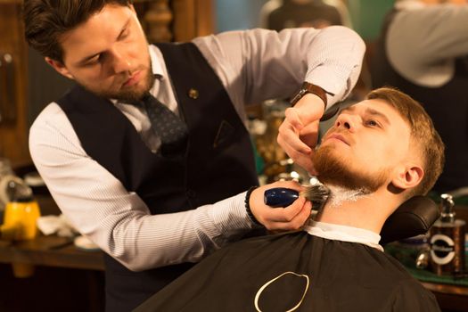 Handsome young bearded man being shaved by a professional barber at the barbershop applying shaving cream on the neck of his customer profession service traditional barbering lifestyle wellness.