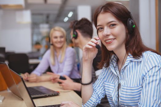 Cheerful helpful female call center operator smiling to the camera, working at the office, copy space. Charming young woman working at customer support service. Happy female at technical support office answering clients calls