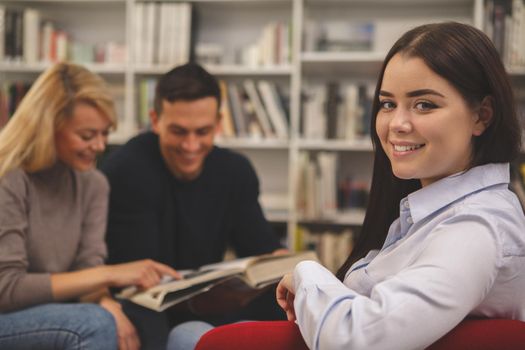 Lovely happy female student smiling to the camera, sitting relaxed at the college library, her classmates reading on the background. Cheerful woman resting in an armchair at the campus library, copy space