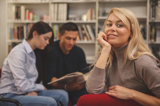 Gorgeous happy blond haired woman smiling to the camera, relaxing at college library with her friends tudying on the background. Lovely female student at the campus. Education, knowledge concept