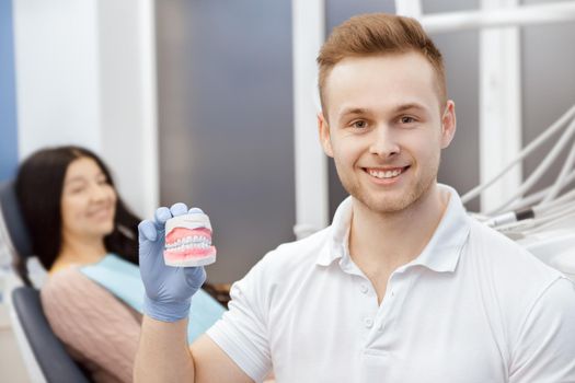 He cares for your health. Cheerful professional dentist smiling to the camera holding dental mold his patient sitting on the back copyspace healthcare medicine professionalism perfect smile concept