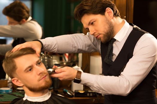 Young handsome professional barber working with his client shaving trimming his beard with a clipper trimmer profession occupation job vintage retro classic lifestyle wellness hairdresser stylist.