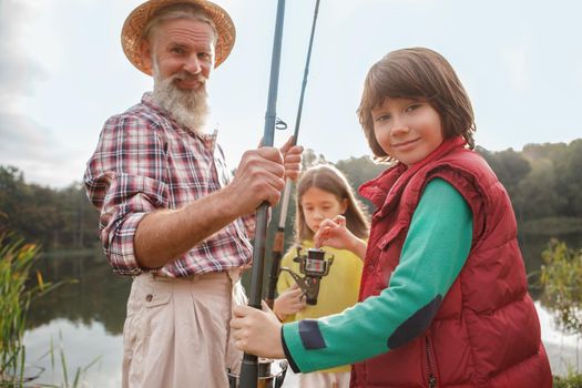 Cute happy young boy smiling to the camera, enjoying fishing with his grandfather and sister on the lake