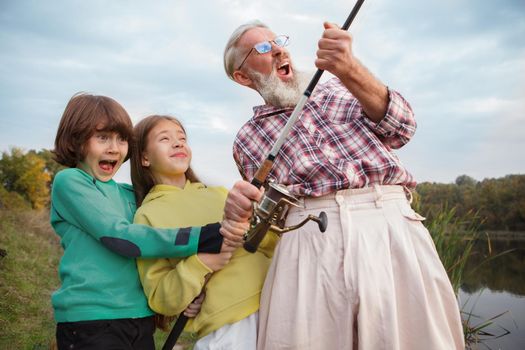 Low angle shot of grandpa and his two grandchildren screaming excitedly, pullin g big fish out of the lake while fishing together