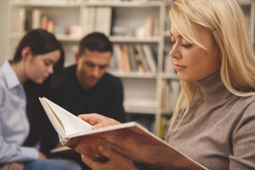 Cropped close up of a beautiful female student reading a book attentively, her college friends studying on the background. Charming woman enjoying reading, copy space. Intelligence, inspiration concept