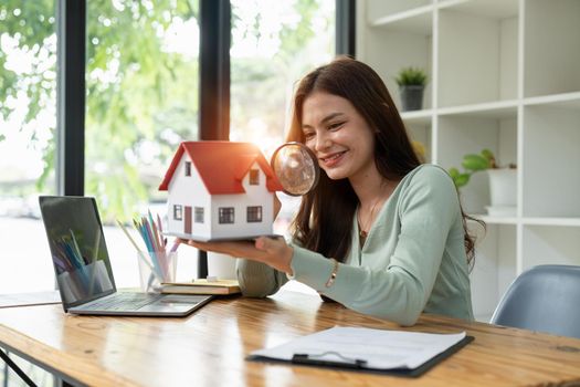 Happy woman hand holding magnifying glass and looking at house model, house selection, real estate concept