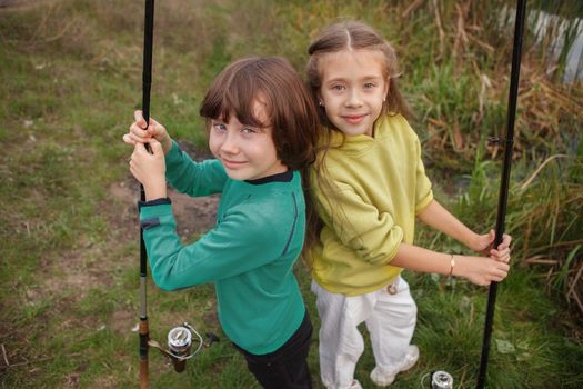 Top view shot of cheerful young boy and girl smiling to the camera, standing back to back holding fishing rods outdoors