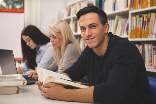 Handsome young man smiling to the camera, while studying with his classmates at the college library. Cheerful male student reading a book, preparing for exams at university. Teamwork, youth concept