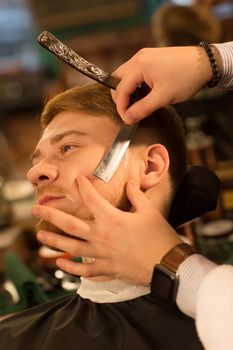 Vertical cropped close up of a young bearded man getting his beard shaved by a professional barber using razor barbering barbershop service lifestyle hipster classic traditional retro blade vintage.