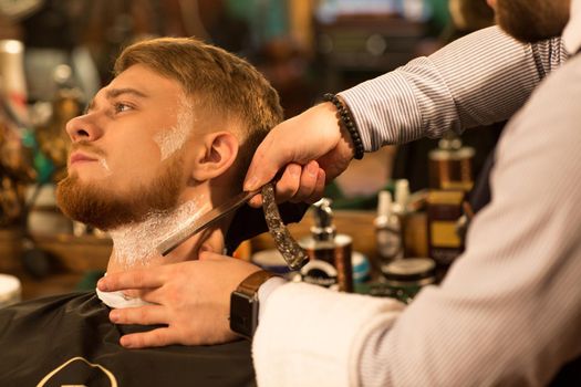 Close up of a young handsome bearded man looking away having his beard shaved by a professional barber with a razor service barbering profession occupation lifestyle hipster classic vintage retro wellness.