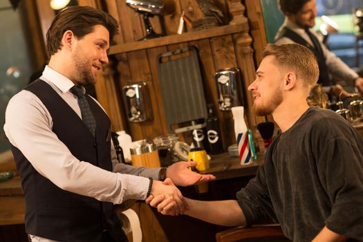 Professional barber shaking hands with his happy male client after giving him a haircut and shaving his beard happiness thankful service communication consumerism hipster lifestyle traditional respect hair salon.