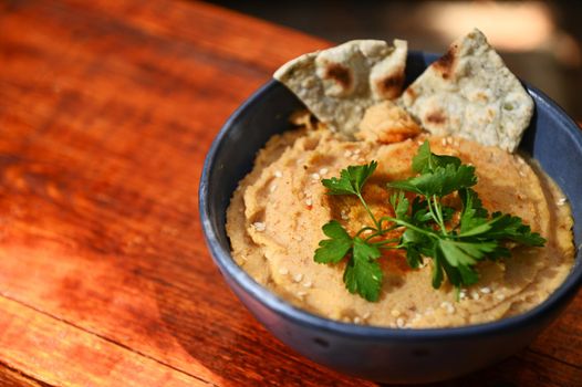 Close-up of a chickpea hummus dip with pita bread, parsley and sprinkled paprika on a blue ceramic bowl, on a rustic wooden table background with copy ad space for text. Still life. Food composition