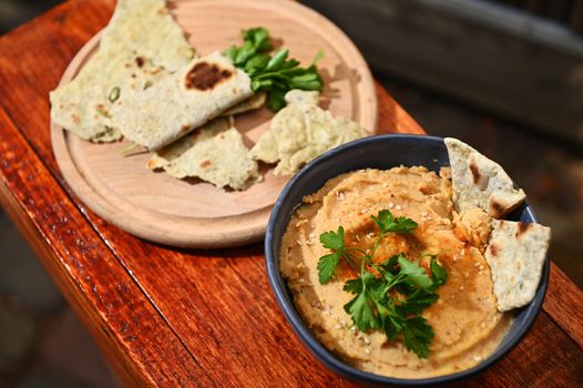 Overhead view of a bowl of a vegetarian chickpea hummus with sprinkled paprika and olive oil and a wooden board with parsley wrapped in pit bread, on a rustic wooden table background