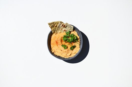 Flat lay of blue ceramic bowl with chickpea hummus, sprinkled with red chili and parsley with pita bread, isolated on white background with copy space for advertising text. Top view. Still life