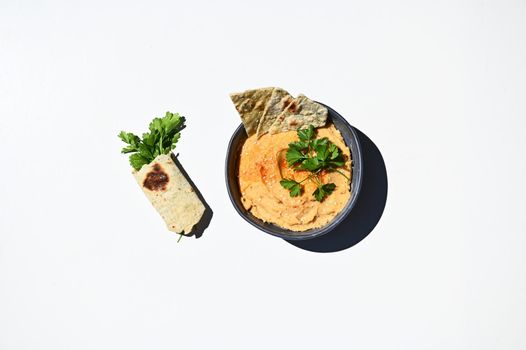 Flat lay composition of blue ceramic bowl with chickpea hummus, sprinkled with red chili and parsley wrapped in pita bread, isolated on white background with copy space for advertising text