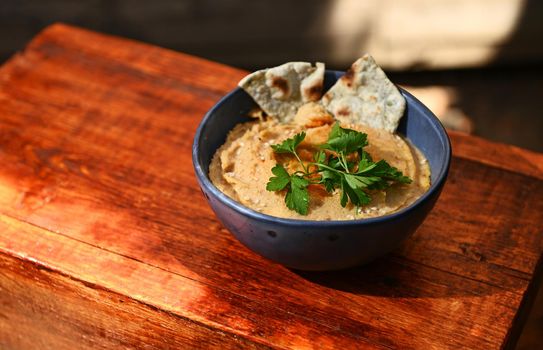Close-up of a chickpea hummus dip with pita bread on a blue ceramic bowl, on a rustic wooden surface with copy ad space for text. Healthy vegetarian food. Food background. Still life