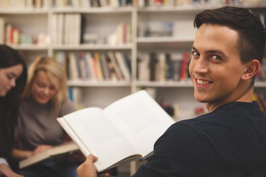 Handsome happy male studenta smiling to the camera over his shoulder, enjoying reading a book at the college library. Cheerful man relaxing at the library reading books, copy space. Education concept